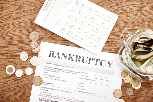 top view of bankruptcy form, coins, calculator and glass jar with dollar banknotes on wooden table