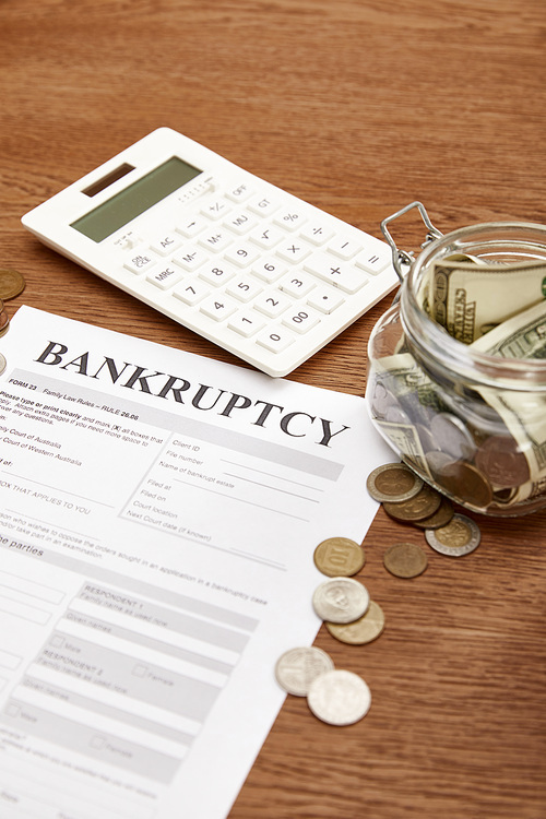 selective focus of bankruptcy form, coins, calculator and glass jar with dollar banknotes on wooden table