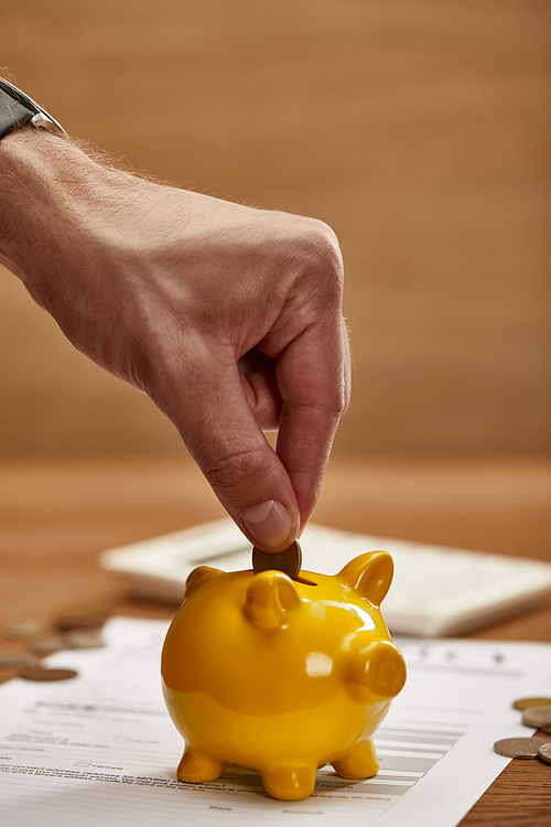 partial view of man putting coin in yellow piggy bank