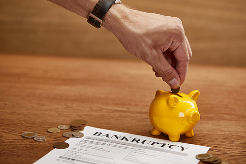 cropped view of man putting coin in yellow piggy bank near bankruptcy form