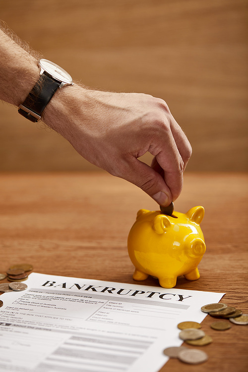 partial view of man putting coin in yellow piggy bank near bankruptcy form