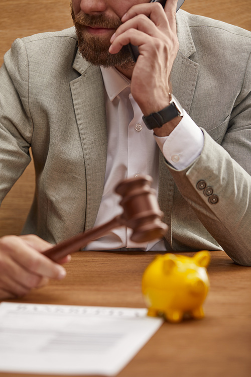 cropped view of businessman in suit talking on smartphone while holding wooden gavel near yellow piggy bank