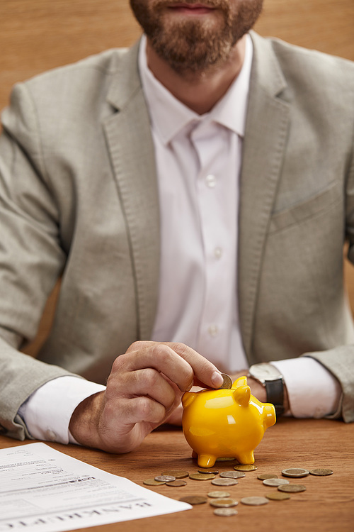 cropped view of businessman in suit putting coin in yellow piggy bank near bankruptcy form