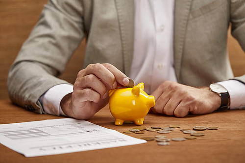 cropped view of businessman in suit putting coin in yellow piggy bank at wooden table