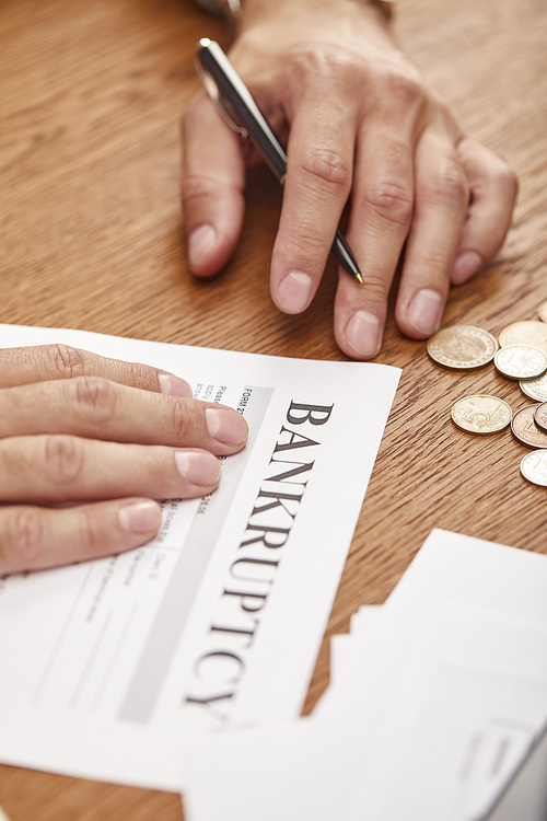 partial view of businessman filling in bankruptcy form at wooden table with coins