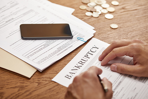 partial view of businessman filling in bankruptcy form at wooden table with smartphone and coins