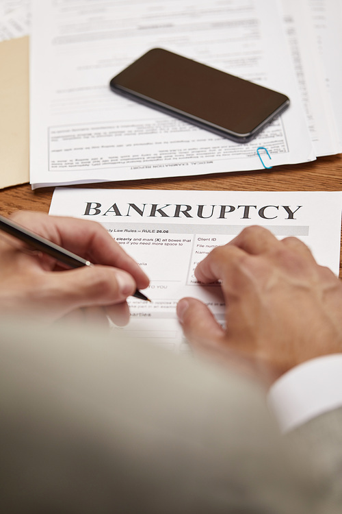 cropped view of businessman filling in bankruptcy form at wooden table with smartphone