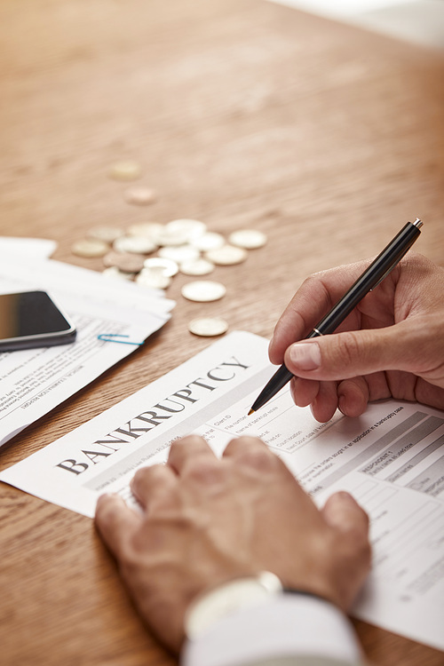 cropped view of businessman in suit  filling in bankruptcy form at wooden table with coins