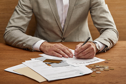 partial view of businessman in suit filling in bankruptcy form at wooden table with documents and credit cards