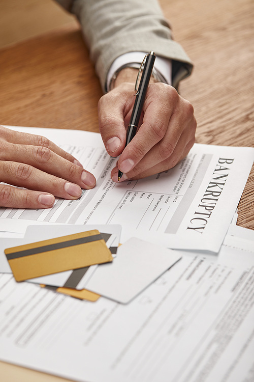 partial view of businessman filling in bankruptcy form at wooden table with documents and credit cards