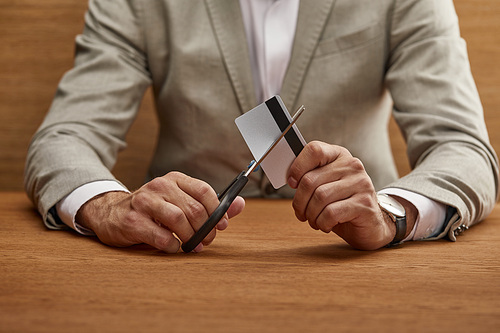 cropped view of businessman in suit cutting credit card with scissors at wooden table