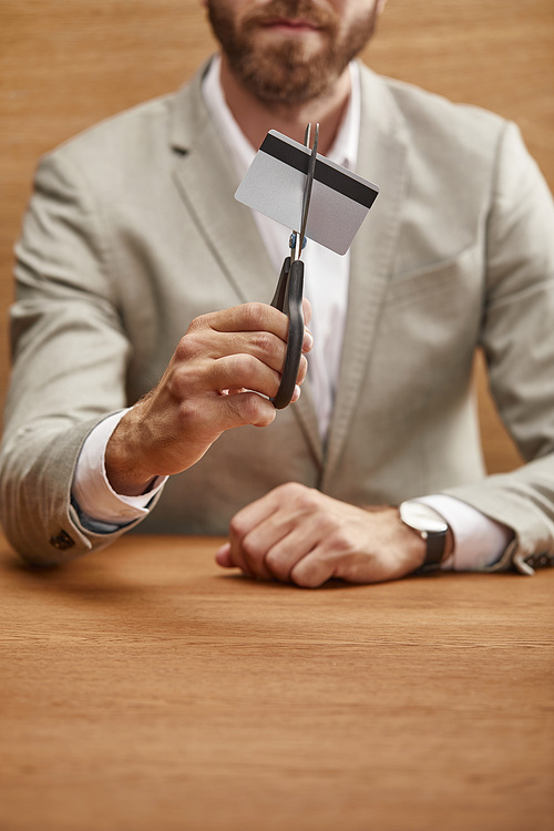 selective focus of bearded businessman in suit holding credit card with scissors at wooden table