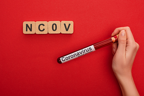 Top view of woman holding test tube with blood sample and coranavirus lettering near wooden cubes with ncov letters on red background
