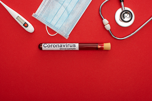Top view of medical mask with thermometer, stethoscope and coronavirus lettering on test tube with blood on red background