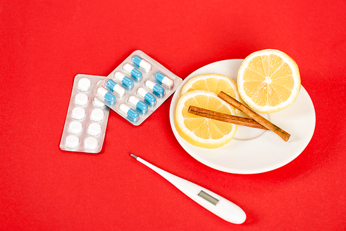 sliced lemons on saucer with cinnamon sticks near pills and digital thermometer on red