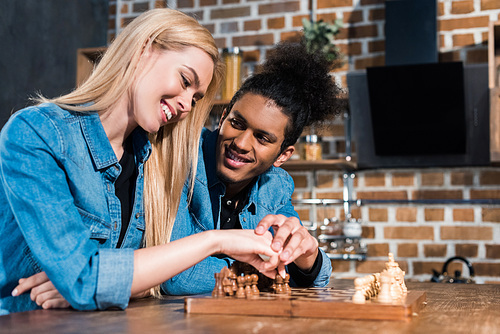 smiling multiethnic young couple playing chess together in kitchen