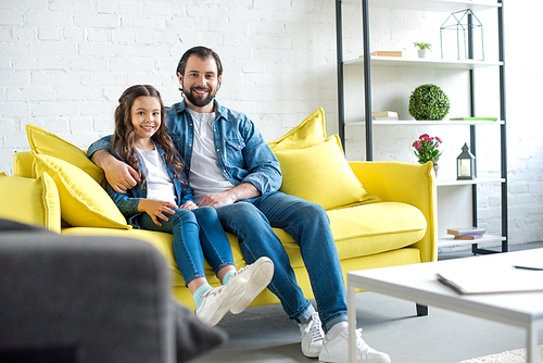 happy father and daughter sitting together on yellow couch and smiling at camera