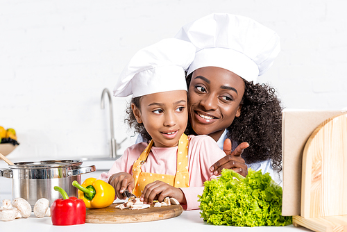 african american mother and daughter in chef hats cutting vegetables and looking into cookbook