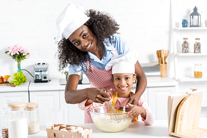 african american mother and daughter in chef hats mixing dough with whisk on kitchen