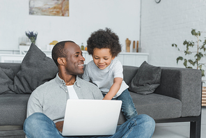 smiling african american father and son using laptop together at home