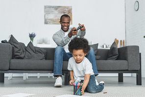 african american father playing video game and son drawing on floor at home