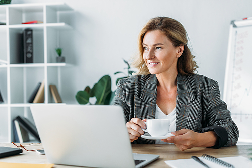 beautiful businesswoman sitting with cup of coffee at table in office and looking away