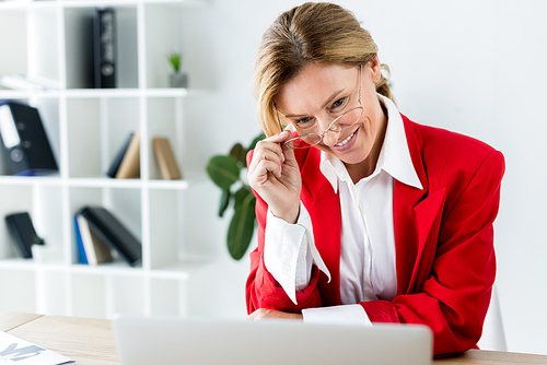 smiling attractive businesswoman touching glasses and looking at laptop in office