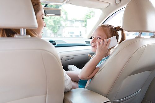 side view of smiling kid talking on smartphone with mother near by in car