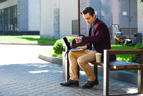 young man using laptop and checking wristwatch while sitting on bench