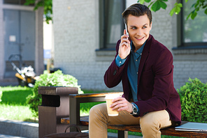 handsome smiling young man holding paper cup and talking by smartphone while sitting on bench