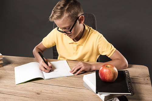 focused school boy in eyeglasses doing homework at table with books, textbooks and apple on grey background