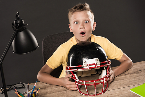 shocked schoolboy sitting with american football helmet at table with eyeglasses, lamp, colour pencils and books on grey background