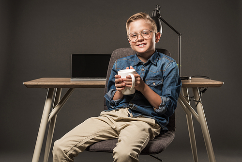 smiling little boy in eyeglasses sitting on chair with coffee cup near table with laptop and lamp on grey background