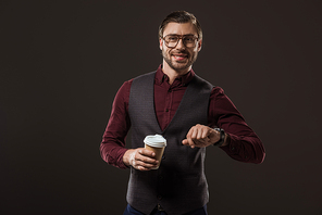 smiling businessman holding paper cup and checking wristwatch isolated on black