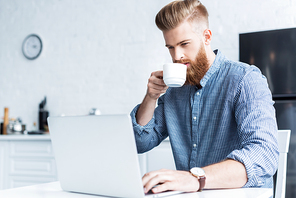 handsome bearded young man drinking coffee and using laptop at home