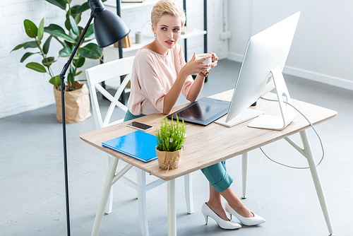 high angle view of female freelancer holding cup of coffee at table with graphic tablet and computer in home office