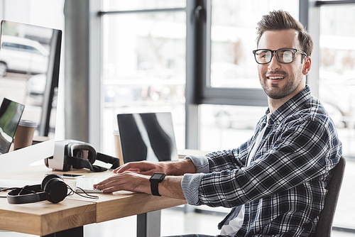 handsome young man in eyeglasses smiling at camera while working with desktop computer and laptop