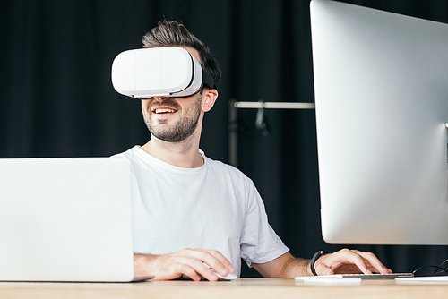 smiling young man in virtual reality headset using desktop computer and laptop