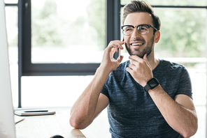 handsome smiling young man in eyeglasses talking by smartphone at workplace