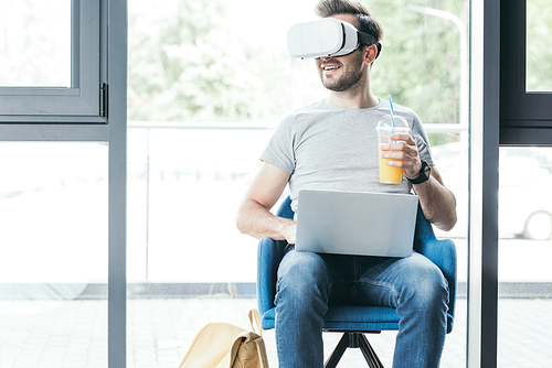 smiling young man holding plastic cup with fresh juice and using virtual reality headset with laptop