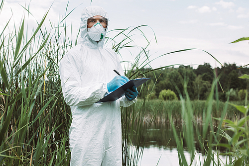 serious male scientist in protective mask and suit writing in clipboard near water outdoors