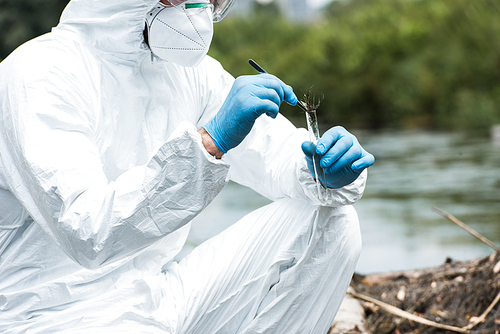 cropped image of male scientist in protective suit and mask putting sample of soil by tweezers in test flask outdoors