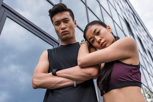 low angle view of serious asian sportsman with crossed arms and sportswoman standing together and  at urban street