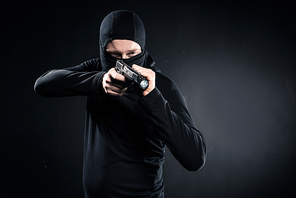 Robber in balaclava aiming with gun on black
