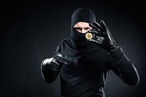 Man in black balaclava holding gun and golden bitcoin