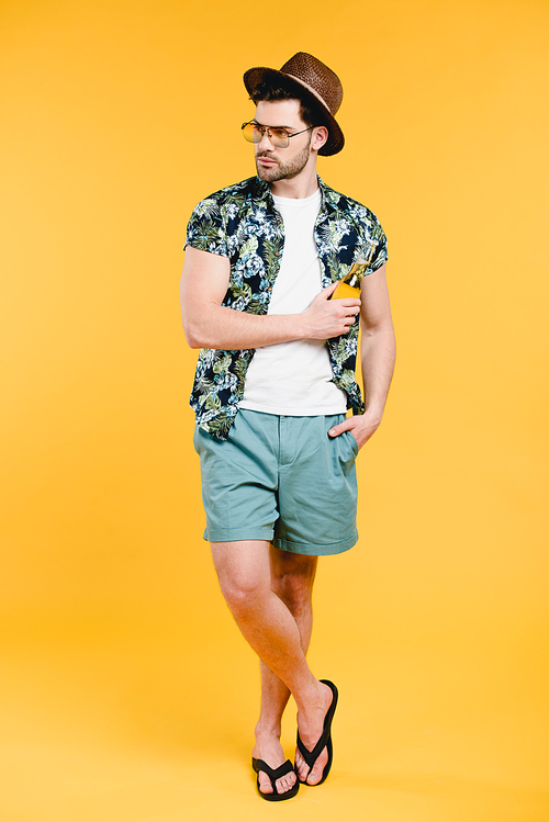 handsome young man in summer outfit holding glass bottle of refreshing beverage and looking away isolated on yellow