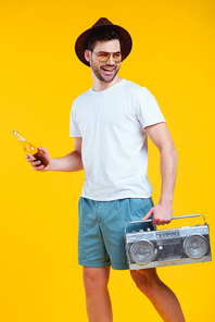 smiling young man in hat and sunglasses holding tape recorder and bottle of summer drink isolated on yellow