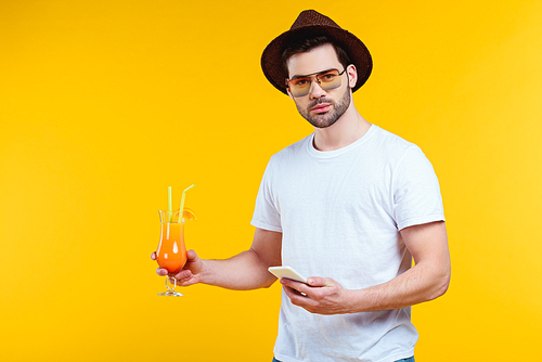 handsome young man  while holding glass of summer cocktail and using smartphone isolated on yellow