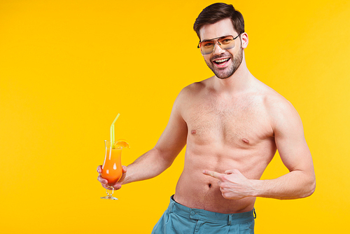 handsome shirtless young man in shorts holding glass of summer cocktail and pointing with finger isolated on yellow