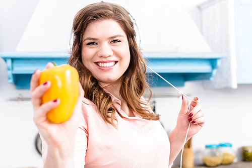 portrait of smiling overweight woman in headphones showing fresh bell pepper in hand in kitchen